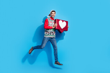 Full size photo of handsome young guy running hold heart icon wear x-mas print vest hat outfit isolated on blue color background
