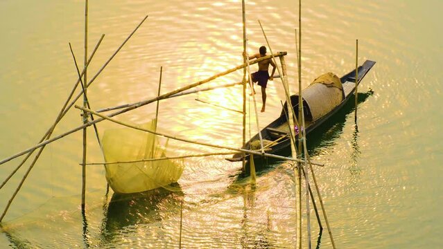 Village boy traps fishing net from boat in river at sunset. Sunlight reflecting and glimmering on river water. Dramatic rural scenery