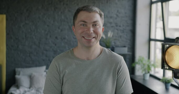 Slow motion potrait of attractive carefree man smiling looking at camera standing in modern bedroom with large window. People and emotion concept.