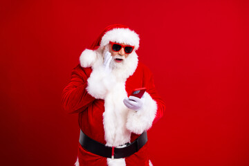 Portrait of astonished positive santa claus in sunglass staring at sale in smartphone hand on beard isolated on red color background