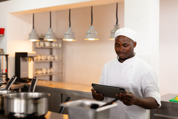 Focused african american male chef standing and using tablet in restaurant kitchen, copy space