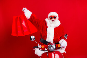Portrait of satisfied santa claus with white beard in sunglass driving scooter raising up shopping bags isolated on red color background