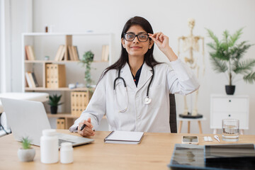 Female general practitioner in medical gown sitting and keeping hand on notebook while working with wireless laptop. Indian woman performing task professionally in field of medicine at office.