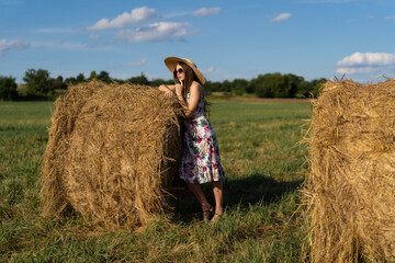 Naklejka na ściany i meble beautiful glamorous girl in a dress and a straw hat on a field near a haystack.
