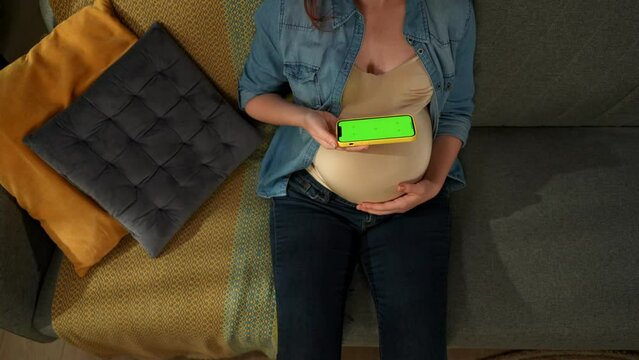 A close up shot shows a pregnant woman sitting on a sofa. She is holding a phone with a green screen in her hands. She examines him. Here can be your advertising. Top view. Medium shot
