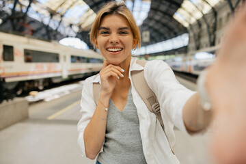 Wonderful stylish girl with happy smile is making selfie and standing on the train g station on...