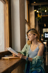 Fototapeta na wymiar Profile portrait of charming cute woman with light hair is reading book while rising in cafe and drinking coffee 