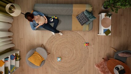 In the shot above, a pregnant woman is sitting on a couch in a room. She is leaning against the back of the sofa, baby toys are lying next to her, she is stroking her belly, smiling, happy.