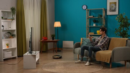 Young man sitting on the sofa in the living room holding smartphone looking at robot cleaner. Smart home concept.