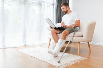 Happy young man with arm in a cast sitting on the couch at home and communicating on a laptop