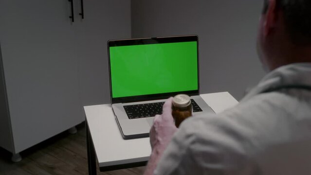 Male doctor sits at table in clinic office looking at laptop with green screen showing jar of medical substance. Profession in medical field. Online conversation with sick patient and taking action