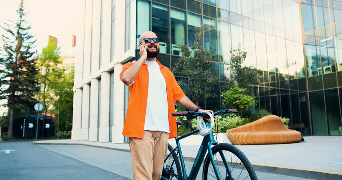Handsome caucasian bearded man in casual clothing talking on a phone walking in the city with bicycle on the way to work near big urban building.
