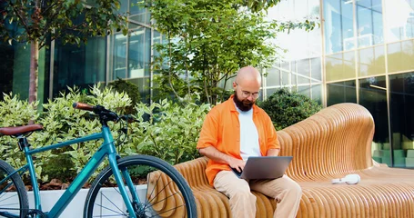 Foto op Canvas Hipster smiling man sitting on bench, typing on laptop computer with bicycle in background. Handsome man working on notebook outdoor near bussines centre. © serg