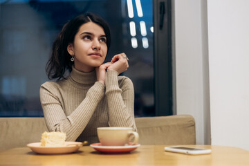 Pretty smiling young woman sitting in cafe, drink cup of coffee hot beverage, eating piece of creamy chocolate cake with nuts on plate, tasting dessert. Chill, relax, lunch 

