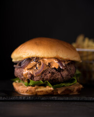 Juicy Homemade burger with lettuce onions, and chipotle sauce in a brioche bun. Dark and moody background. French Fries in background.