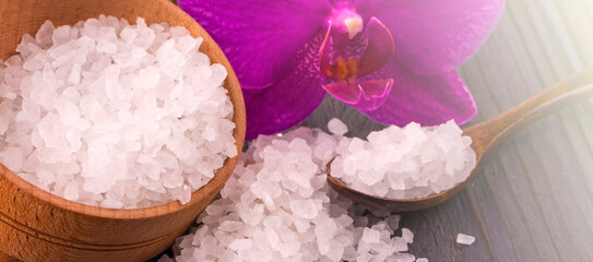 White bath salt in a wooden bowl with a spoon and an orchid on wooden table