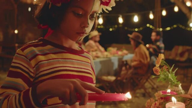 Tilt up of Hispanic little girl with half painted face lighting candle at altar during Dia de Muertos family celebration in backyard at night