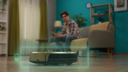 Man sitting on the couch and holding smartphone, turning on robot vacuum cleaner using app, battery infographic visible over cleaner. Smart house concept.