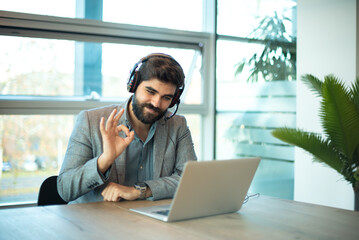 Multicultural businessman wearing call center agent headset showing OK sign with fingers, smiling friendly gesturing excellent symbol to laptop's screen. - obrazy, fototapety, plakaty