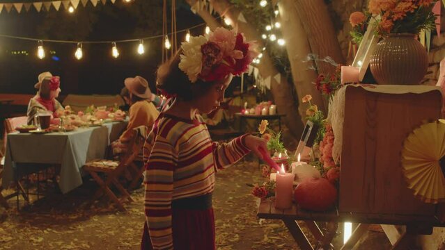 Medium shot of Latin girl in colorful striped sweater and flower wreath lighting candle at altar during Day of the Dead family celebration in backyard in evening