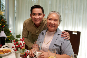 Portrait of cheerful Vietnamese man posing with his mother at dinner table