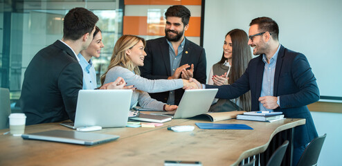 Welcome to our team! Smiling CEO female entrepreneur shaking hands with cheerful businessman while business partners coworkers clapping hands and celebrating in boardroom.