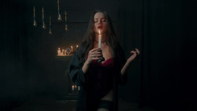 Young sexy witch or vampire with a candle in her hands in a dark room in slow motion. Sexy young white girl with a candle in her hands in a dark room