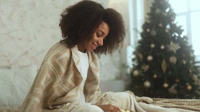Portrait of happy positive young curly woman with popcorn while watching comedy movie show serial enjoying leisure time during holidays sitting on bed at home near Christmas tree indoors