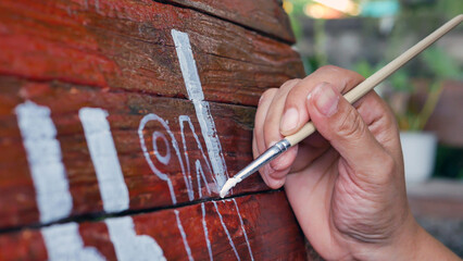 Close-up of Hands of Young woman painter creating art use a paintbrush to draw white lettering designs on a wooden coffee shop sign.outdoor activities,People doing activities. - obrazy, fototapety, plakaty
