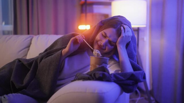 Portrait of sad young woman dealing with stress by eating food sitting on couch at home Upset brunette female wrapped in blanket crying and eating ice cream indoors at late night Mental heath problem