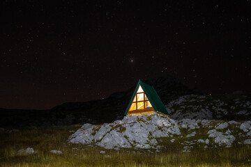 Beautiful small a-frame mountain shelter house with glass wall lit inside at night with stars in the sky