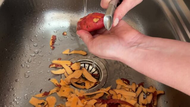 a woman peels a sweet potato fruit with a vegetable peeler over a metal sink under running water from a tap in the kitchen, cooking