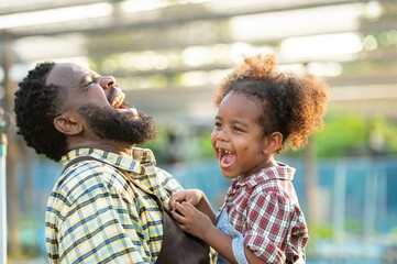 Happy african black parents dad father with daughter child on piggyback teasing fun in garden greenhouse. Black daughter kiss cheek dad and neck riding in vegetable greenhouse garden in light sunset - Powered by Adobe