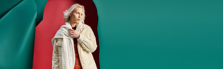 young blonde woman in winter jacket adjusting knitted scarf on red with turquoise backdrop, banner