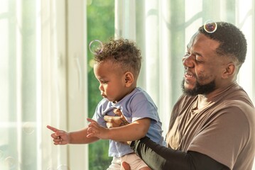 Happy african black parents dad father throw baby son on piggyback and neck riding teasing fun near window. Black baby son and daddy enjoy teasing kiss cheek and neck riding at window light - obrazy, fototapety, plakaty