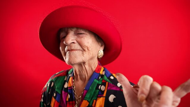 Fisheye portrait of funny elderly mature woman, 80s, wearing red hat giving shaka hand gesture isolated on red background. Concept of old rock and roll person