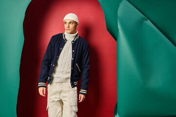 stylish young man in beanie, white sweater and bomber jacket on ripped red with turquoise backdrop