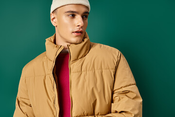 handsome man in beanie hat and stylish puffer jacket on turquoise background, winter trends