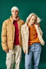 fashionable man in winter puffer jacket hugging woman and posing together on turquoise background
