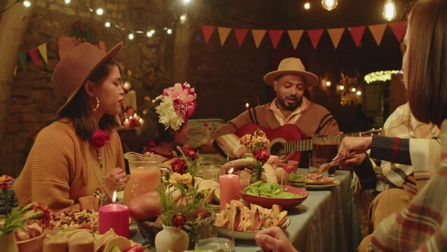 Waist up of modern big Mexican family of five having festive dinner in backyard on Day of the Dead, playing guitar, eating and talking