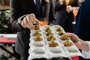 torso of caucasian man in suit reaching for mini crab cake canape garnished with herb sauce at...