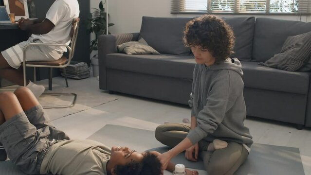Tilt up shot of Caucasian mother taking care of her African American son lying on floor resting from doing physical exercises with his father working in background