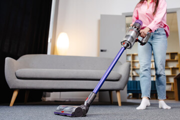 Young woman uses cordless vacuum cleaner to clean home carpet. Modern easy cleaning