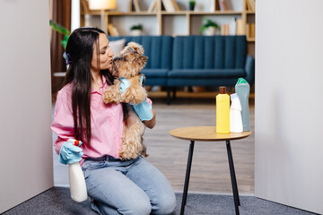 A young pretty woman wearing rubber gloves holds her small dog in her arms and a cleaning agent in the other hand