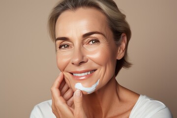 Close-up of middle-aged Caucasian woman touching her face to apply moisturizer. Smiling face of adult grey-haired lady with daily cream, facial cosmetics. Skin care. Grey background, copy space.
