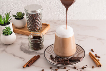 A jet of hot chocolate is poured into a large glass of coffee, cappuccino or cinnamon latte. Front view marble background. A chic dessert.
