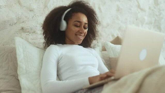 Charming curly woman relaxing at home listening music or working online browsing products in internet store reading news checking email or social media profile on laptop computer laying on bed indoors