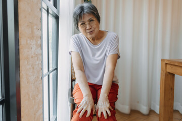 Asian old woman holding and touching knees pain joint, sitting next to window with painful face looking at camera, bad symptom health care.