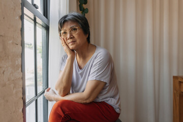 Asian old woman sitting on chair, resting chin on hand and looking out the window, missing and waiting family and thinking something sad, living alone.