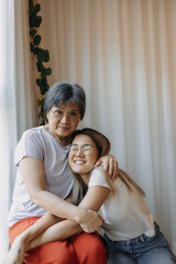 Happy Asian Thai mother and daughter looking at camera, smiling with love, cuddle hugging while mom sitting on chair at home, good family relationship.